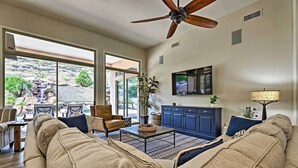 A spacious living room with a large sofa, a coffee table, and a TV mounted on the wall. There is a ceiling fan, a window with a view of a rocky landscape, and a blue cabinet. - Patio, Playground! Glendale Family Gem Near Golf (Glendale)