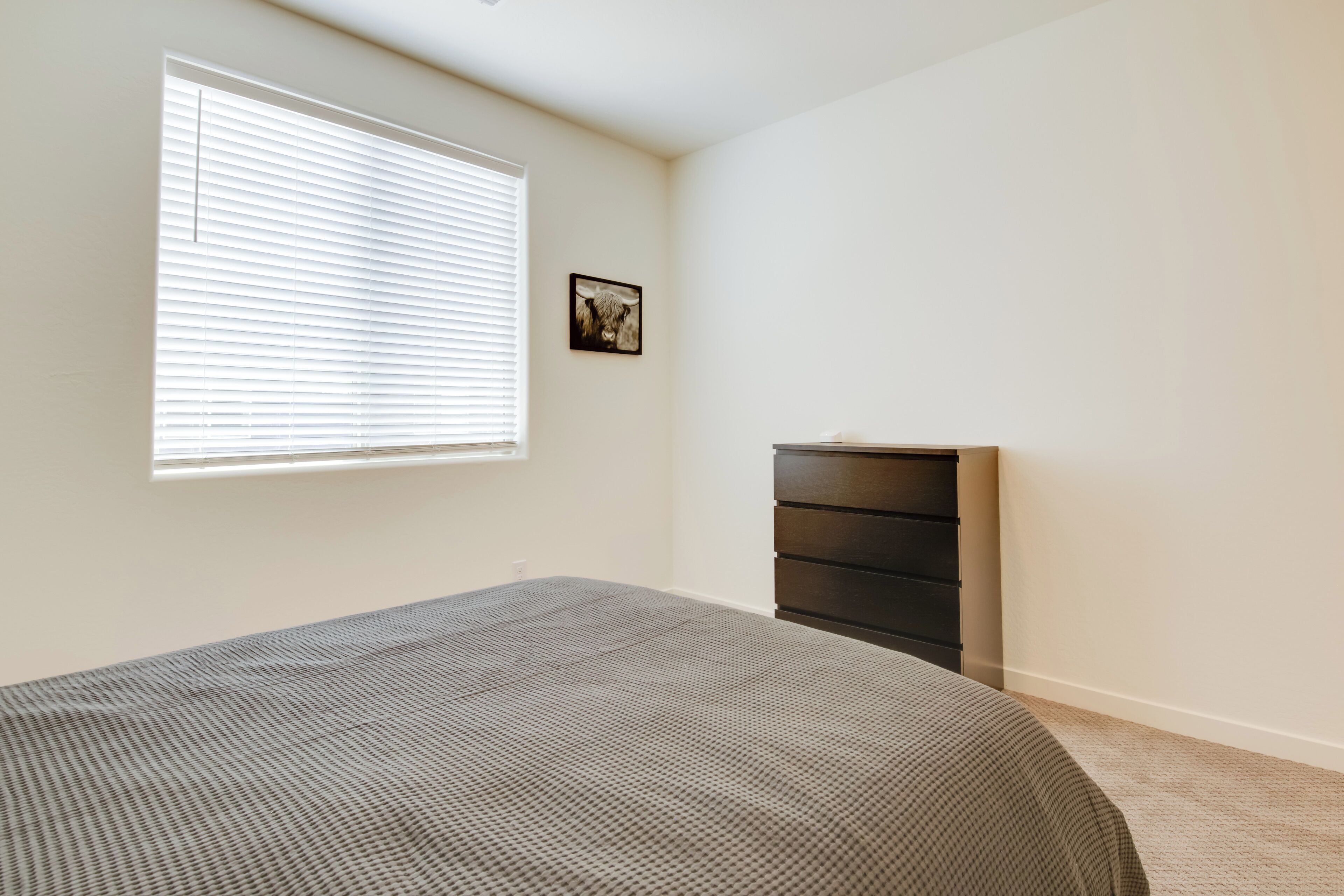 Ein Schlafzimmer mit Bett, Kommode, einem Fenster mit Jalousien und einem Wandbild.