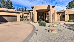 A residential property with a curved driveway, a central entrance with a covered porch, and a landscaped garden with rocks and shrubs. - Lavish Estate w/ Sports Court & Home Theater! (Scottsdale)