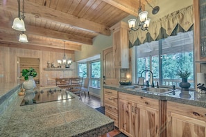 Una cocina con gabinetes de madera, encimeras de granito y una isla grande. Hay una zona de comedor con sillas y una mesa, y una ventana con persianas.