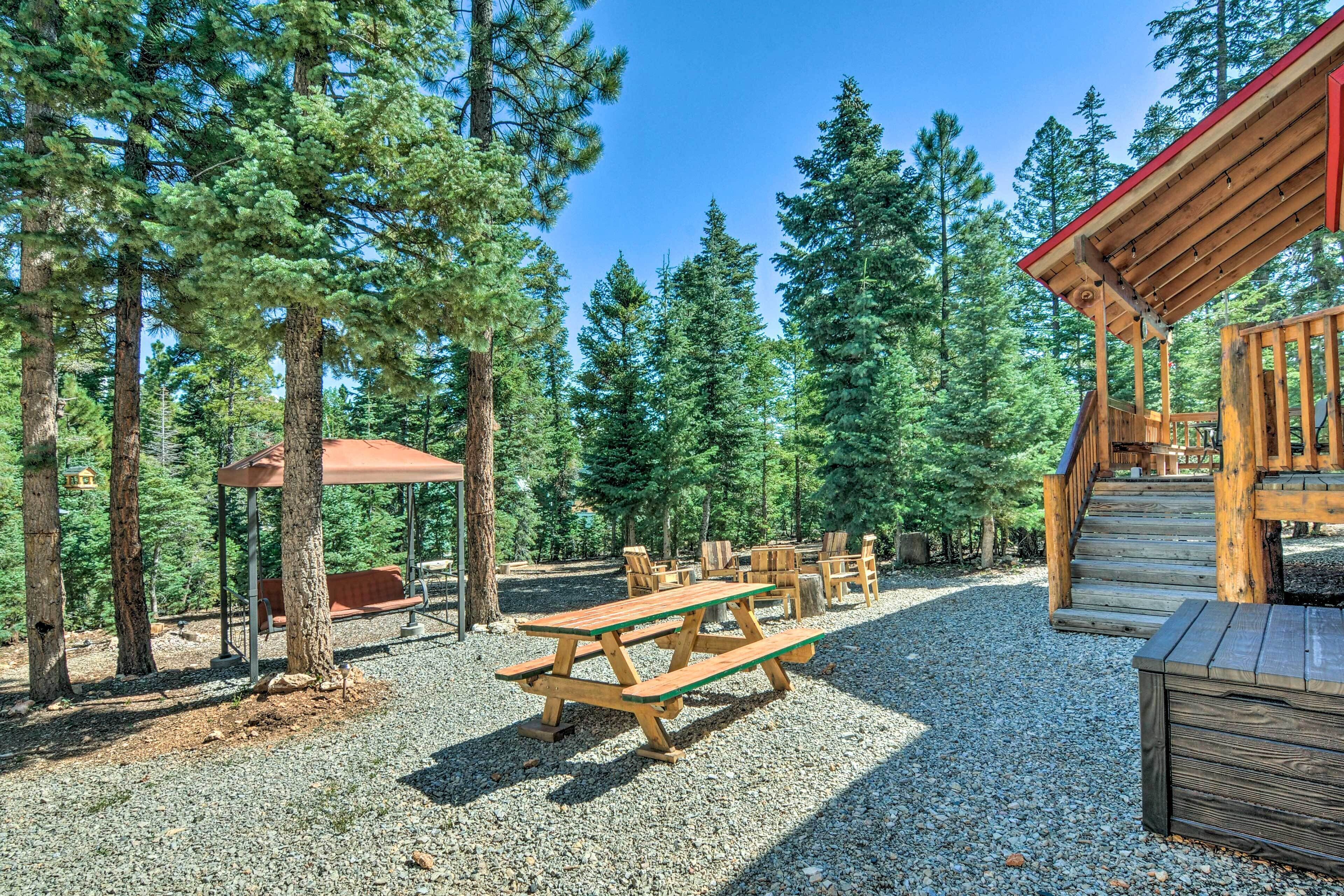 A wooden picnic table and benches in a forested area with a covered seating area and a staircase leading to a building.
