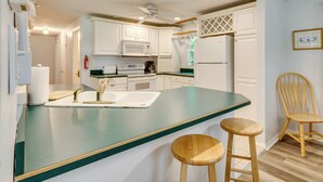 Une cuisine avec des armoires blanches, un plan de travail vert, un ventilateur de plafond et deux tabourets en bois.
