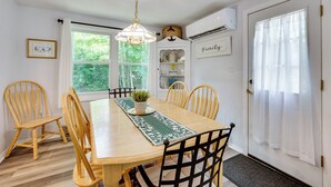 Un coin repas avec une table en bois, six chaises, un lustre et une vue sur la verdure environnante.