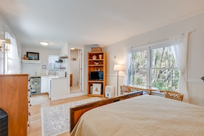 A bedroom with a large bed, a wooden dresser, a desk with a chair, a television, and a kitchen area in the background. - Quaint Smoky Mountain Cabin Rental w/ Patio (Highlands)