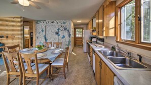 A kitchen with wooden cabinets, a dining table with chairs, a ceiling fan, and a window with a view of trees. - Lakefront Home With Deck, Dock, & Water Access! (Liberty)