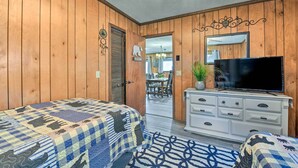 Une pièce aux murs recouverts de lambris en bois, un lit avec une couvre-lit à motifs, une commode sur laquelle est posée une télévision, et un coin repas à l’arrière-plan.