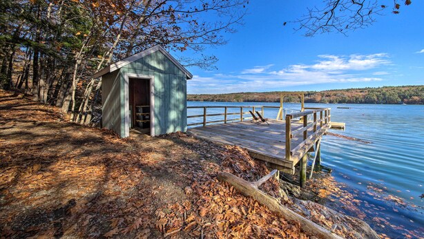 A wooden dock extending into a calm lake, with a small shed nearby. - Riverfront Coastal Escape With Deep Water Dock! (Wiscasset)