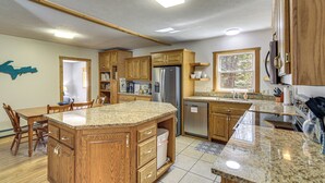 A kitchen with wooden cabinets, granite countertops, a stainless steel refrigerator, and a dining area with chairs. - Powell Lake 'UP Badger Lodge Bnb' w/ Shared Dock (Wetmore)