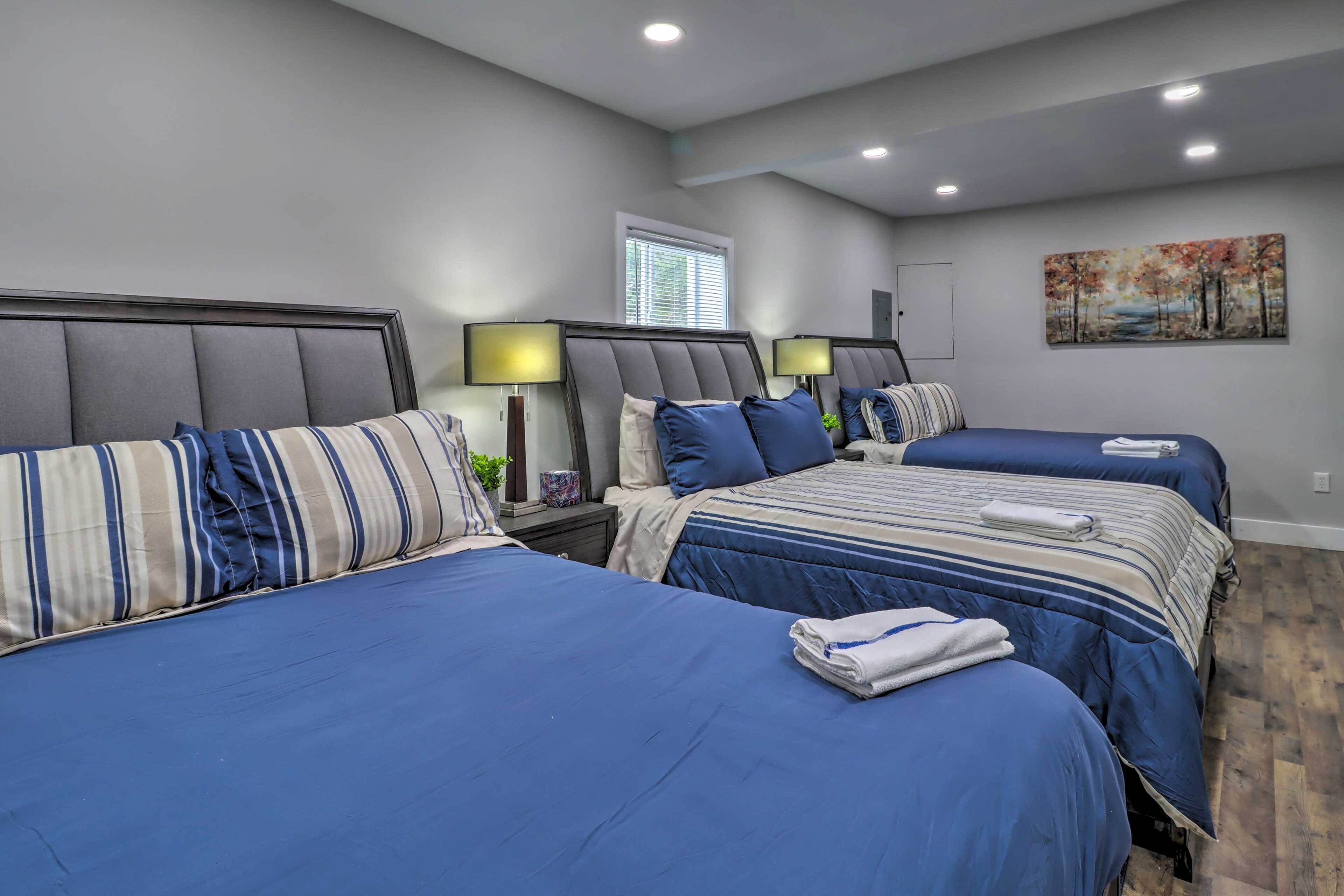 Quarto de hotel com três camas, cada uma com roupa de cama azul e almofadas listradas. Há mesas de cabeceira com luminárias e um quadro na parede.