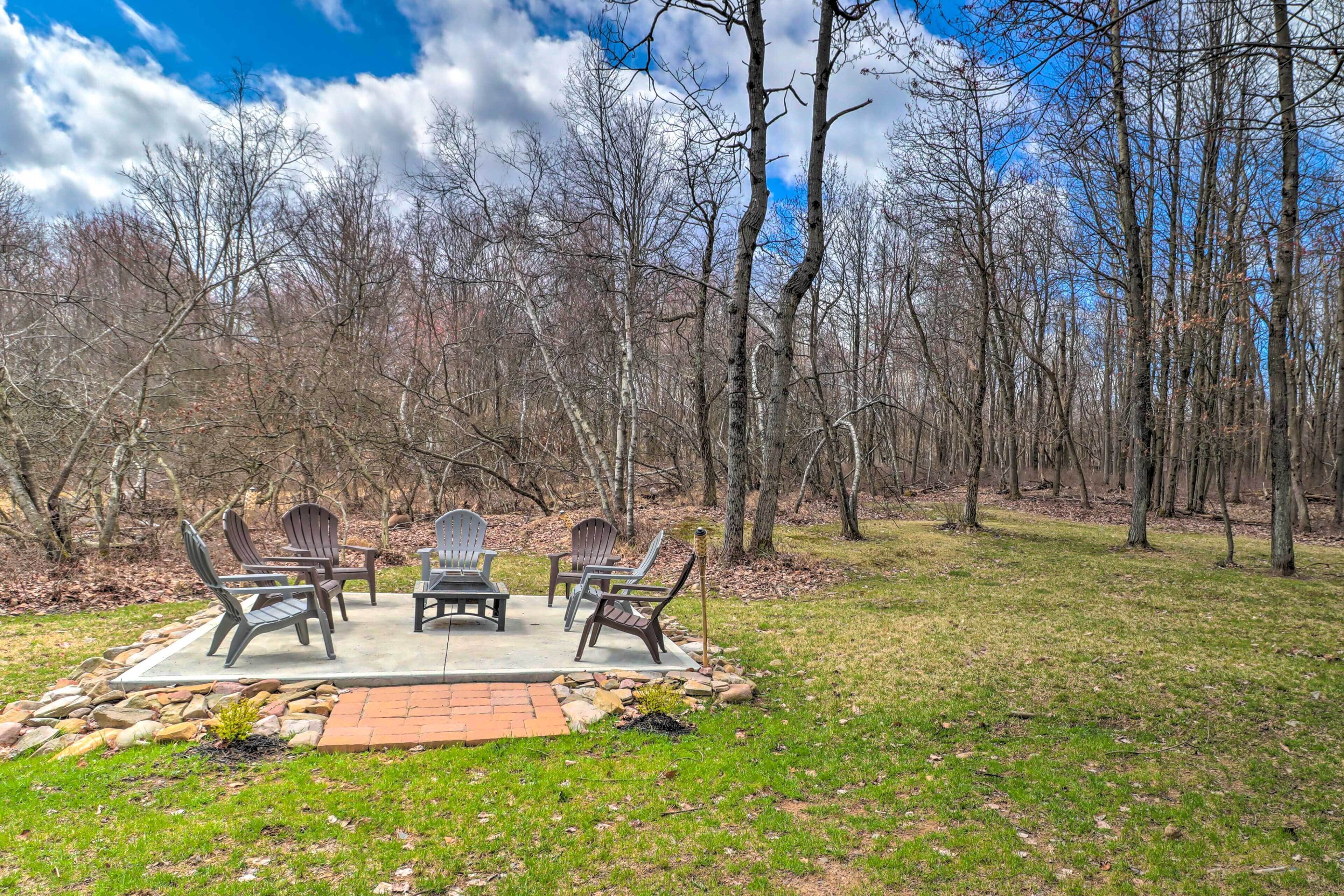 An outdoor seating area with Adirondack chairs and a fire pit, surrounded by a wooded landscape.