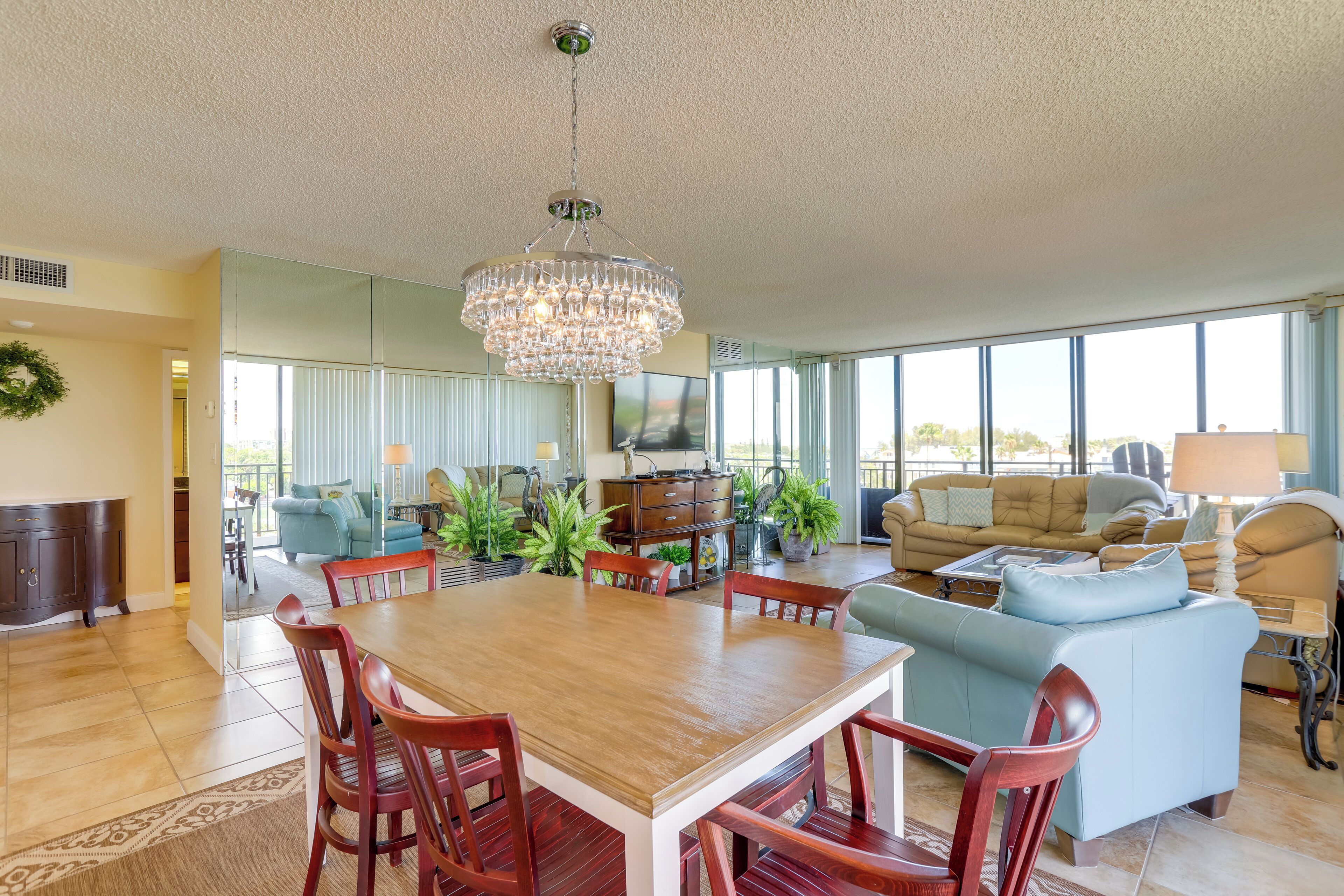 A spacious living room with a wooden dining table, red chairs, a chandelier, and a large window offering a view of the outdoors.