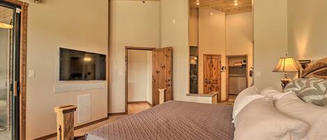 A bedroom with a wooden ceiling fan, a bed with a gray quilt, a TV mounted on the wall, and a wooden wardrobe.