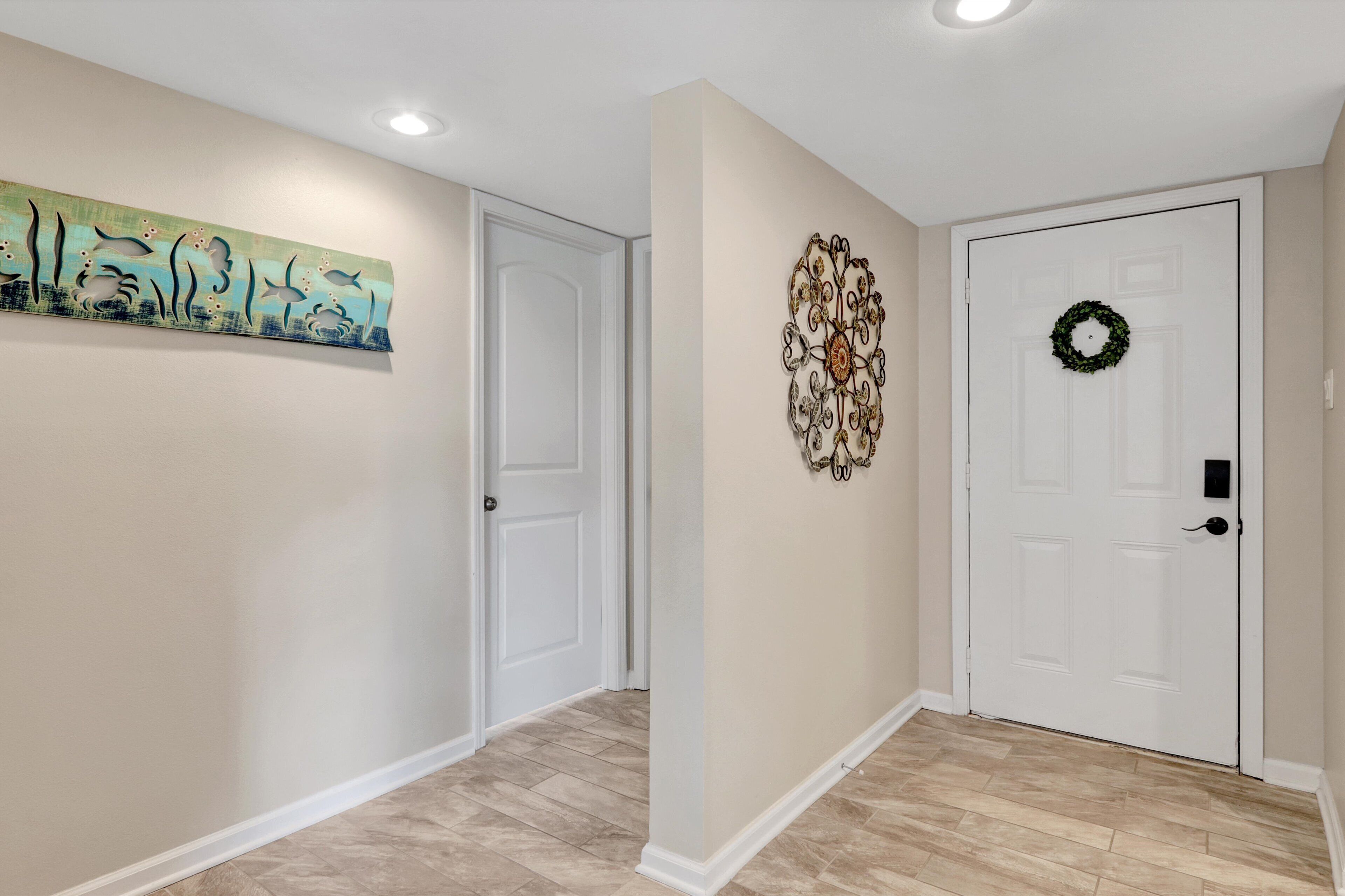 A hallway with beige walls, a wooden floor, a white door with a wreath, and a decorative metal piece on the wall.