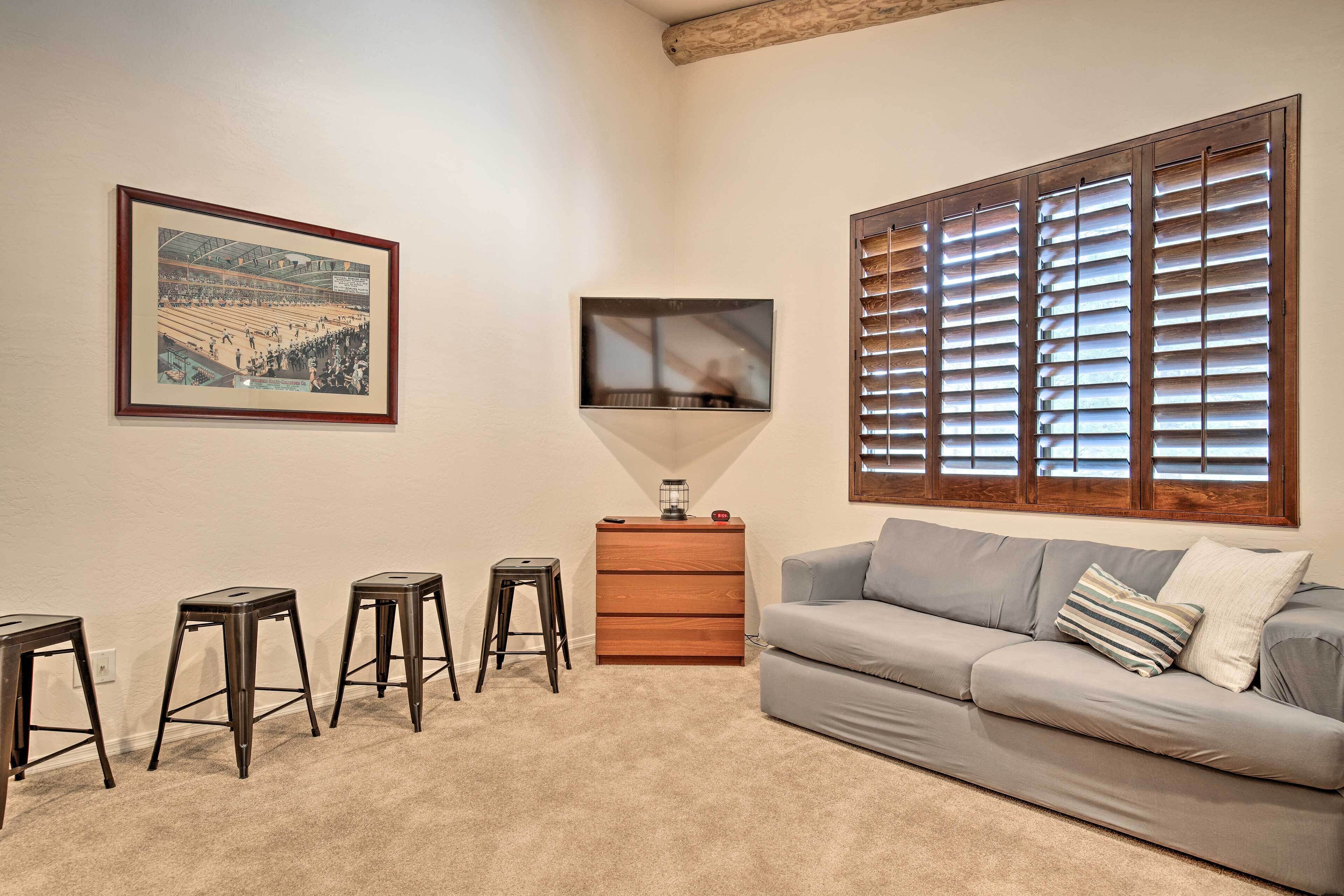 A living room with a grey sofa, a wooden side table with a lamp, a framed picture on the wall, and a window with blinds.