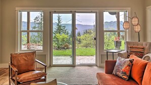 A spacious living room with a large sliding glass door leading to a garden. There is a leather armchair and a sofa with cushions.