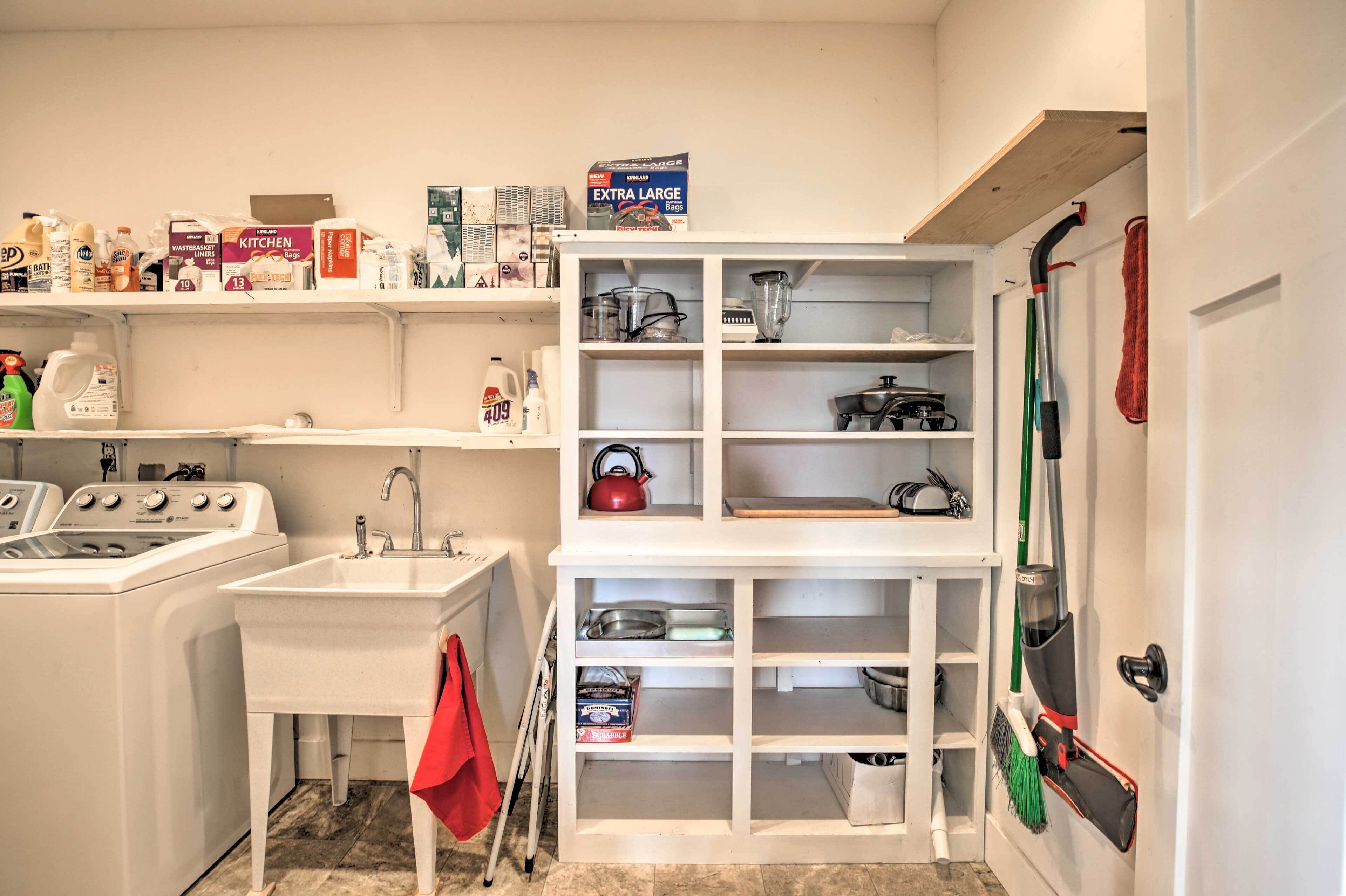 A laundry room with a washing machine, sink, and shelves stocked with cleaning supplies and kitchenware.
