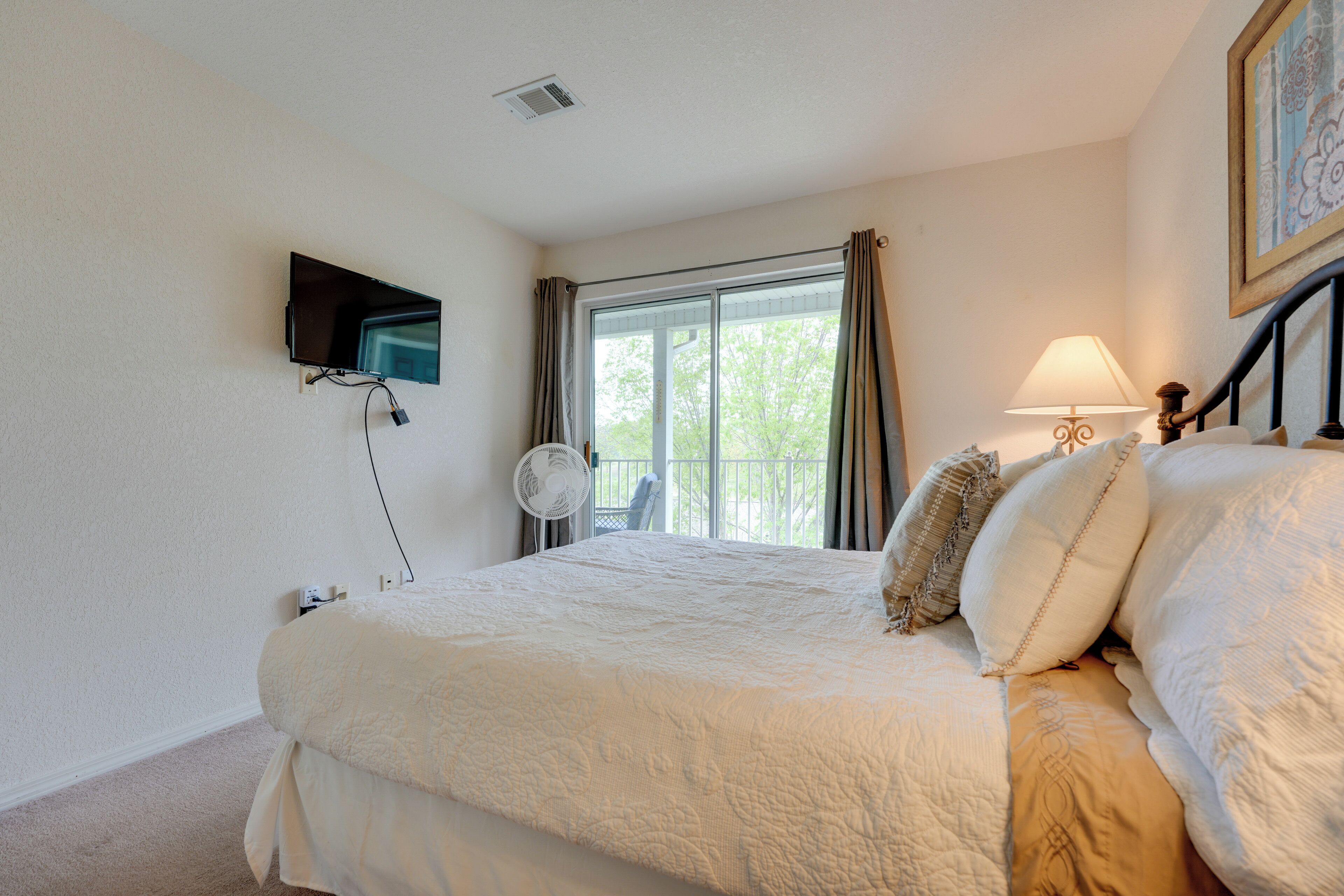 A bedroom with a bed, a TV mounted on the wall, a window with curtains, and a bedside lamp.