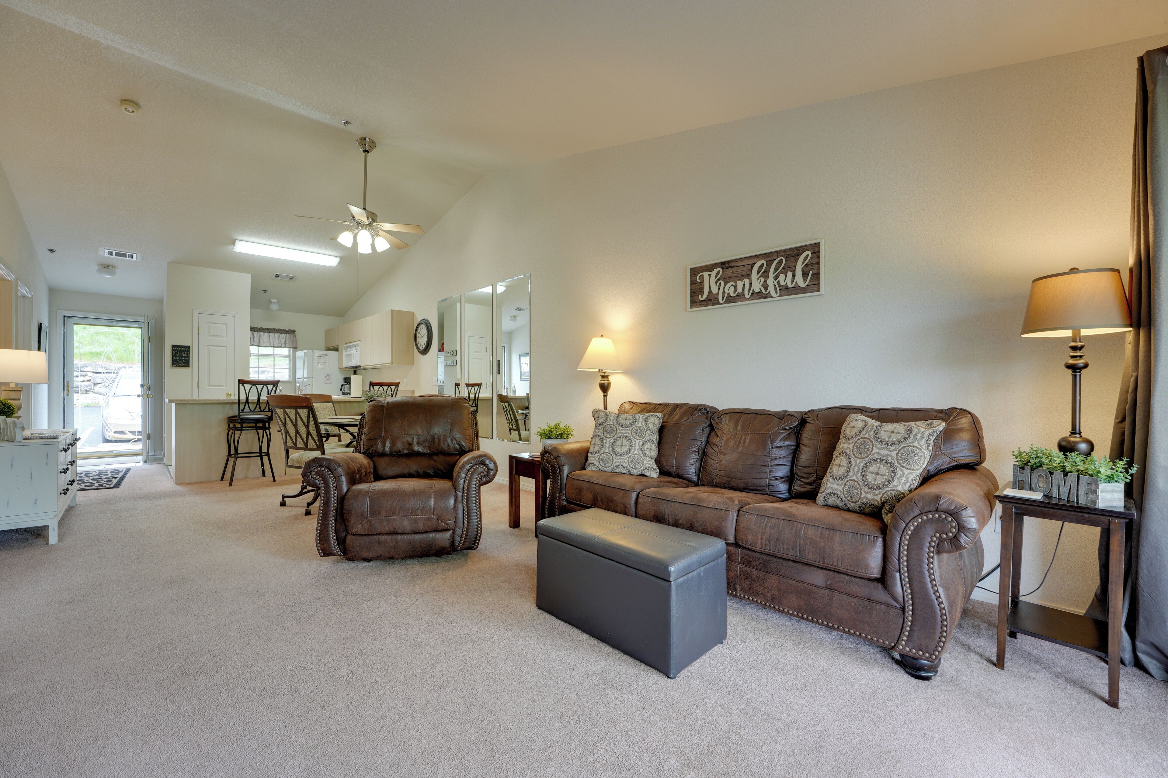 A spacious living room with a brown leather sofa, a matching armchair, and a grey ottoman. A wooden side table with a lamp and a small plant is visible. The room has a large mirror, a clock, and a 'Thankful' sign on the wall.
