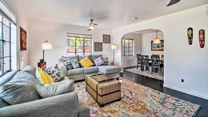 A modern living room with a grey sofa, a wicker ottoman, and a colorful rug. There are two pendant lights, a ceiling fan, and a dining area visible in the background. - Sun-lit Phoenix Gem: Backyard Entertainment Patio! (Phoenix)