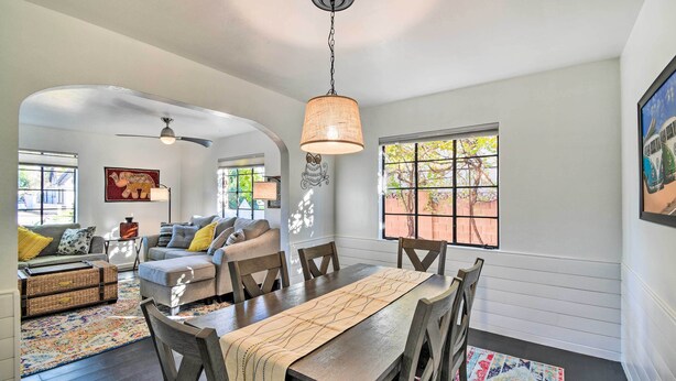 A dining area with a wooden table, chairs, and a hanging light fixture. There is a window with a view of greenery outside. - Sun-lit Phoenix Gem: Backyard Entertainment Patio! (Phoenix)