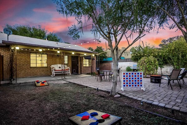 A backyard with a brick house, a wooden bench, a table, chairs, and a game board. - Sun-lit Phoenix Gem: Backyard Entertainment Patio! (Phoenix)