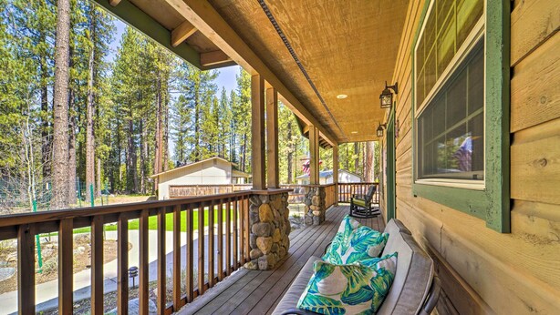 Une terrasse en bois avec un pilier en pierre, un banc agrémenté d’un coussin à motifs de feuilles, et une vue sur une zone boisée.