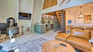 A spacious living area with a high ceiling, a wooden staircase, a cozy sofa, and a central fireplace. - Wilmington Chalet w/ Hot Tub < 8 Mi to Mount Snow! (Wilmington)