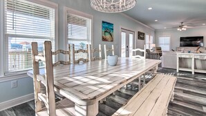 A dining area with a wooden table and chairs, a chandelier, and a view through large windows. - Luxe Stilted Home w/ Kayaks: Walk to Beach (Dauphin Island)