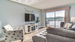 Un salon doté d’un grand téléviseur à écran plat, d’un meuble de rangement blanc, d’un canapé gris et d’un balcon offrant une vue sur l’eau.