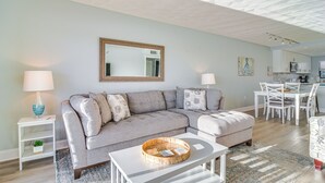 Un salon moderne avec un canapé d’angle gris, une table basse blanche et un coin repas comprenant des chaises et une table blanches.