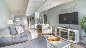 Un salon avec un canapé gris, un fauteuil à motifs, un téléviseur à écran plat et un meuble de rangement blanc pour les appareils multimédias.