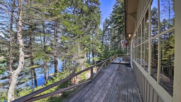 A wooden deck with a railing overlooking a forested area and a body of water. - Cozy Cottage on Cat Cove w/ Wraparound Deck! (Stonington)
