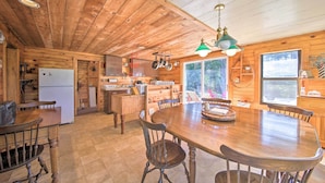 A wooden cabin interior with a large dining table, chairs, a kitchen area, and a refrigerator. - Cozy Cottage on Cat Cove w/ Wraparound Deck! (Stonington)