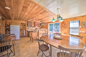 Interior de una cabaña de madera con una gran mesa de comedor, sillas, una zona de cocina y un refrigerador.