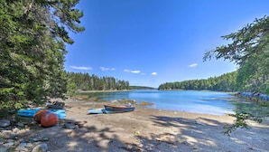 A serene lakeside setting with a boat on the shore, surrounded by dense forest. - Cozy Cottage on Cat Cove w/ Wraparound Deck! (Stonington)