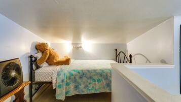 A small, single-bed room with a bed, a chair, a fan, and a wall-mounted light fixture.