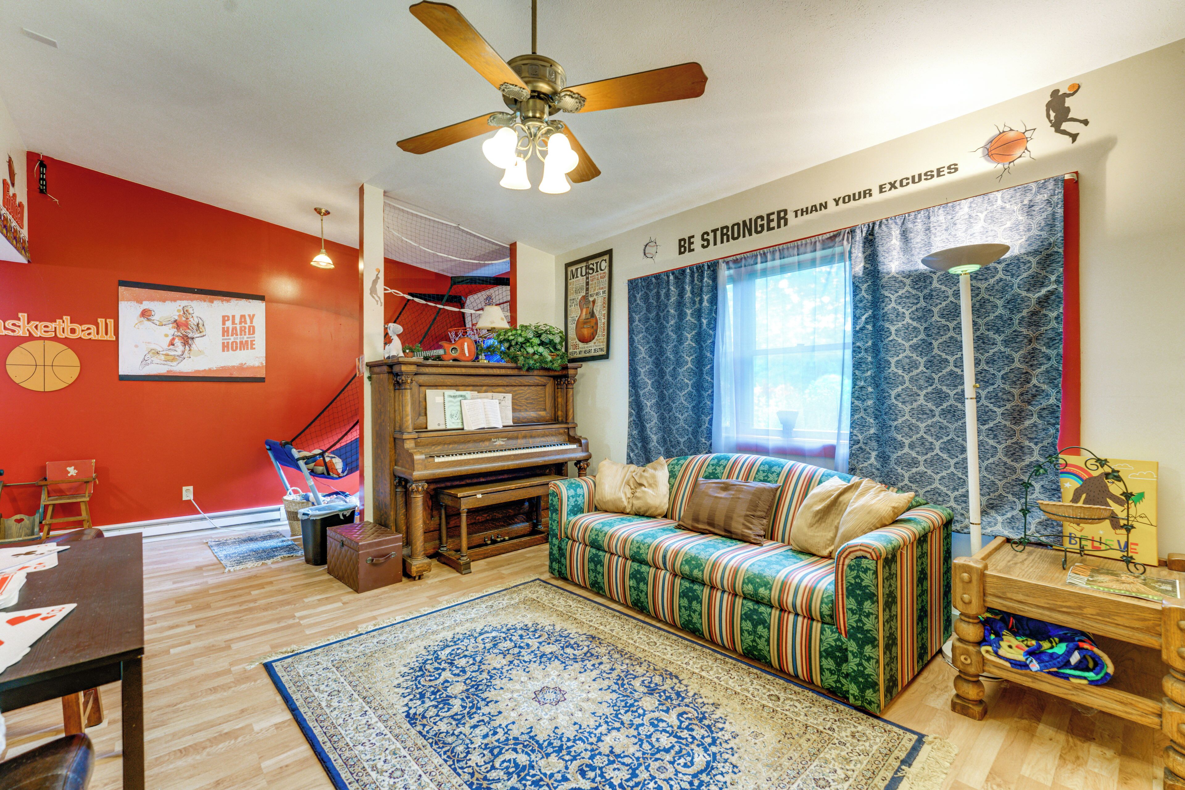 A living room with a red wall, a piano, a sofa, a patterned rug, and a ceiling fan.