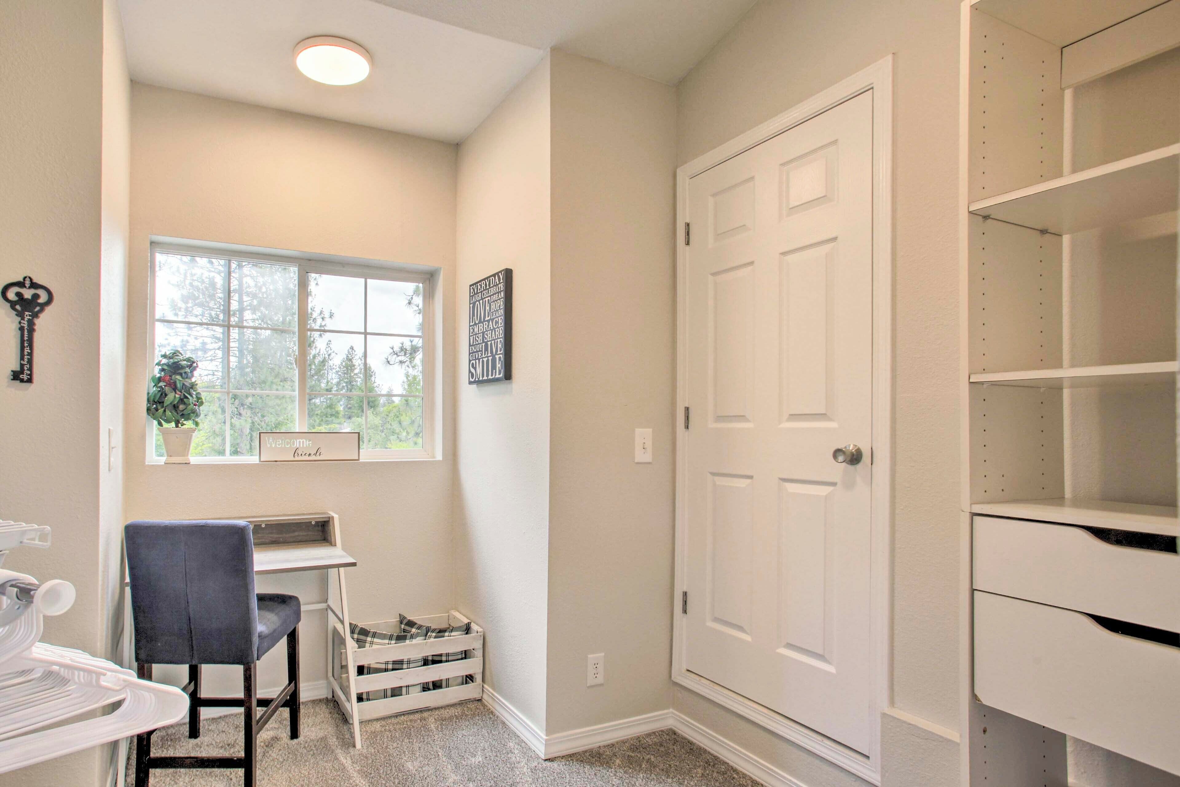 A room with a desk, chair, and a window with a view of trees. There is a white door and a shelf unit.