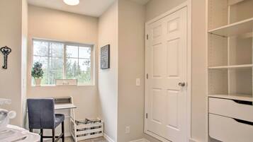 A room with a desk, chair, and a window with a view of trees. There is a white door and a shelf unit.