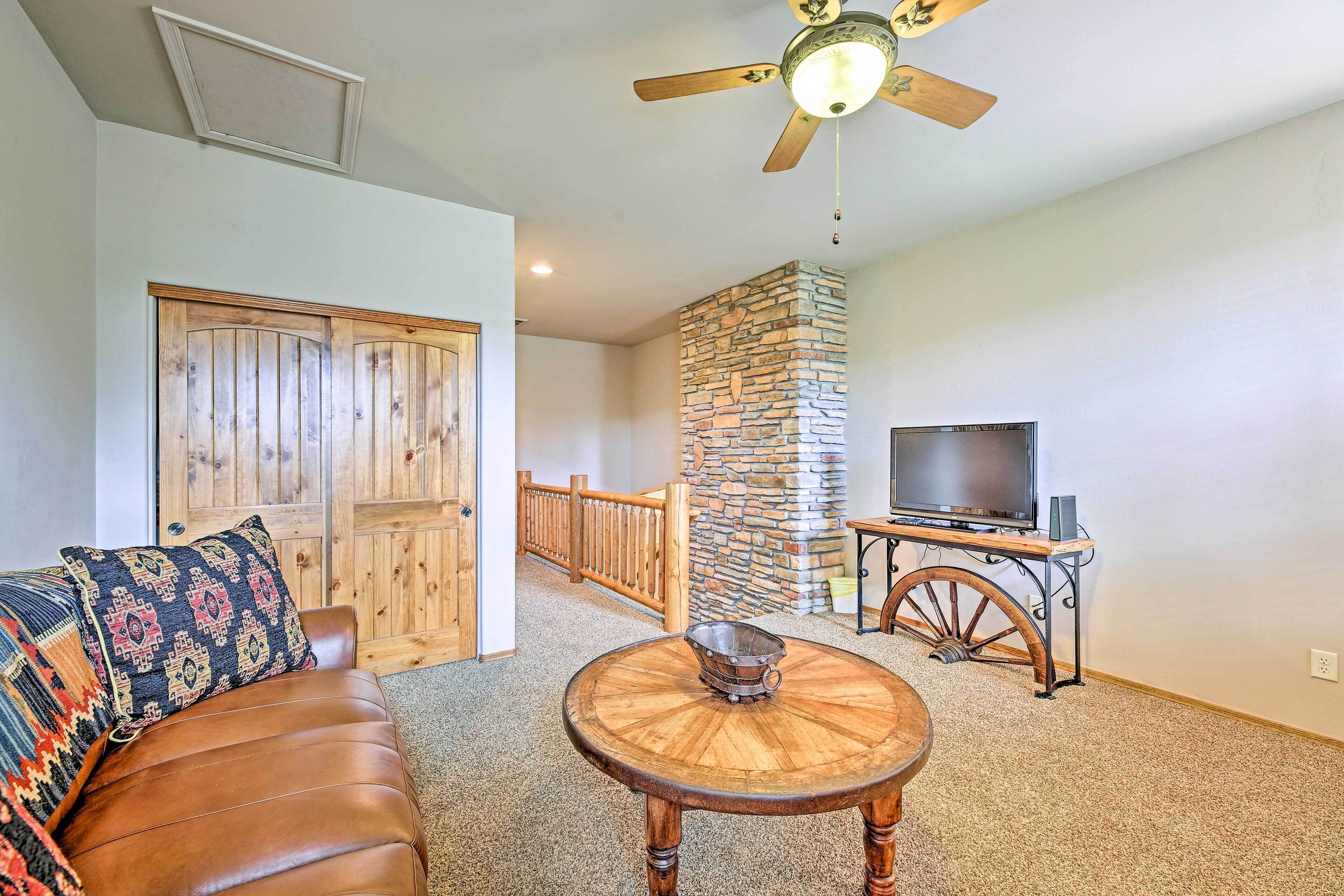 A living room with a brown leather sofa, a round wooden coffee table, a stone fireplace, a television on a wooden cart, and a ceiling fan.