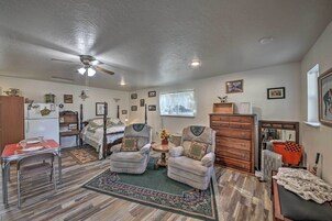 Una habitación acogedora con cama, dos sillones, un aparador de madera, una mesita con un jarrón y un ventilador de techo.