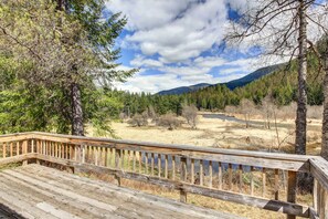 A wooden deck with a view of a river and forested mountains. - Angler's Dream Remote Escape on Bull River! (Noxon)