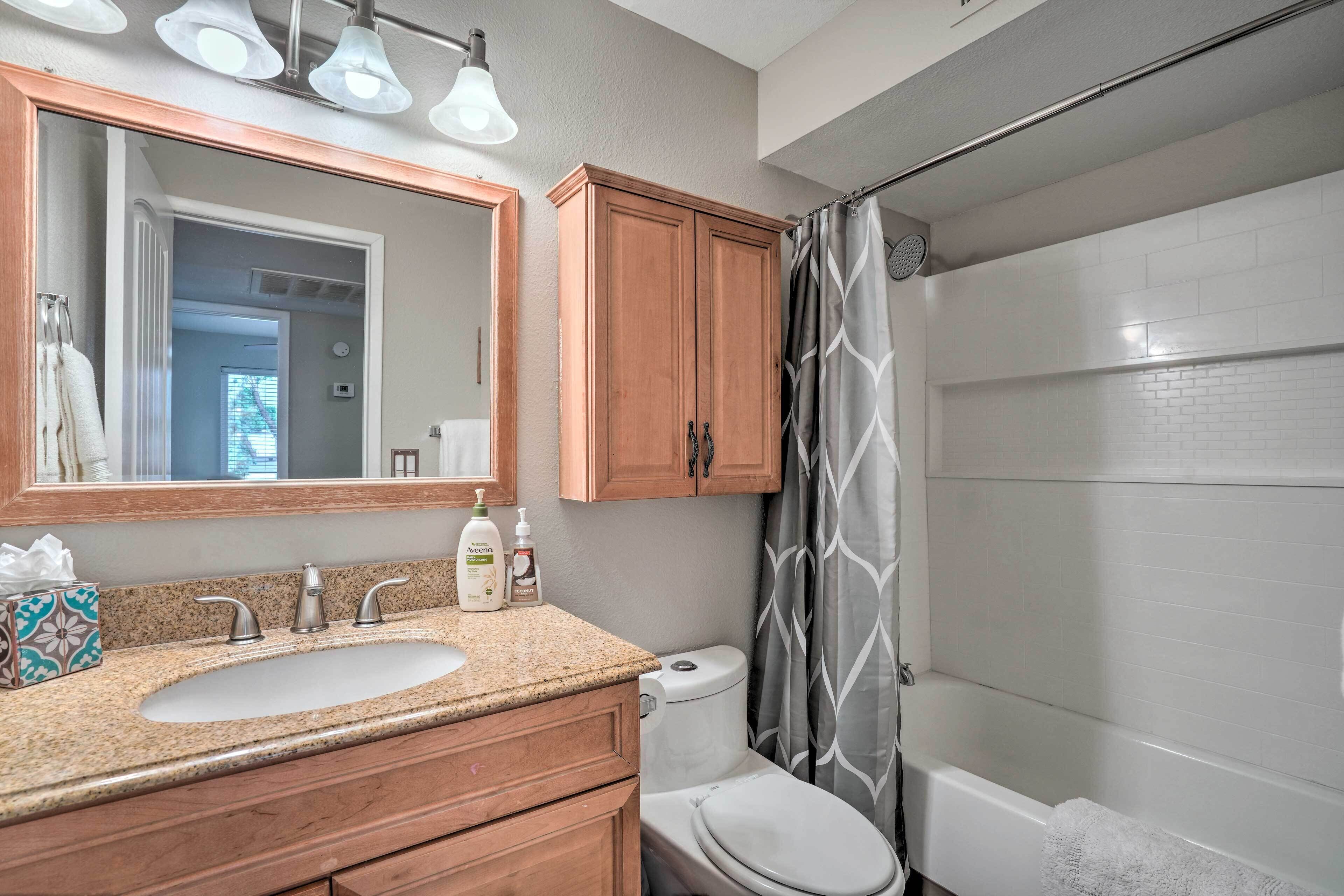 Une salle de bain avec un lavabo, des toilettes et une douche. Un miroir est placé au-dessus du lavabo et il y a un rideau de douche orné d’un motif de feuilles.