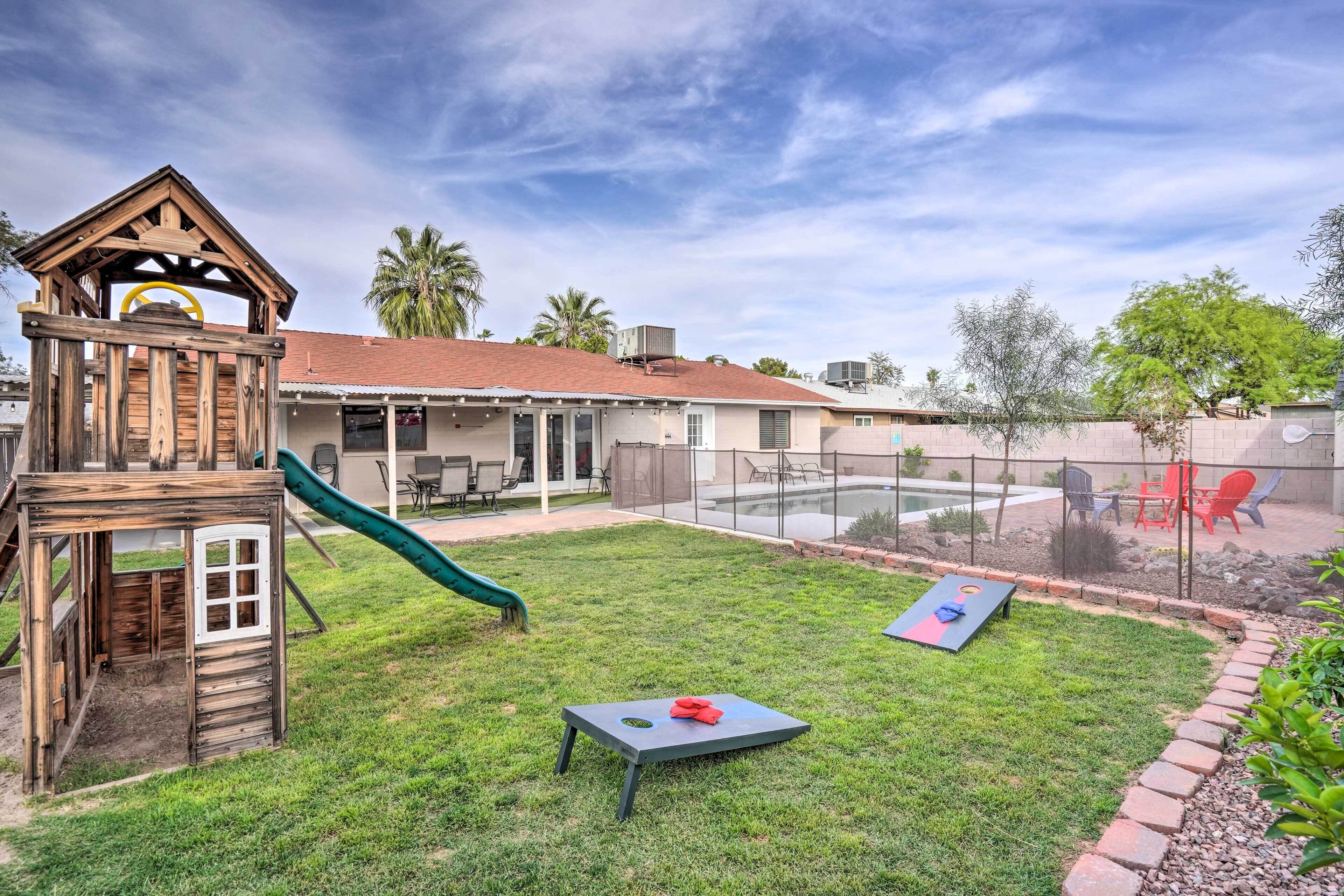 A backyard with a wooden playhouse, a slide, a table with a red object, and a pool area.