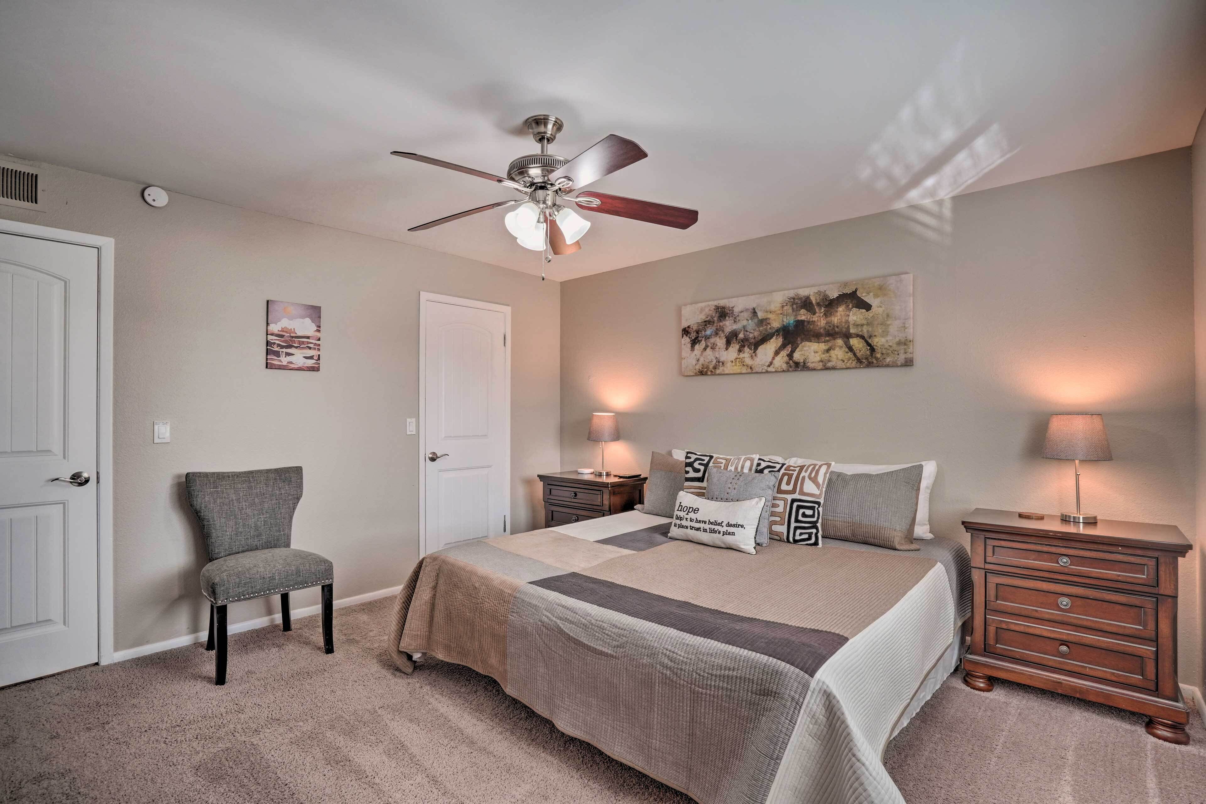 A bedroom with a bed, a chair, a nightstand, a lamp, a ceiling fan, and artwork on the walls.