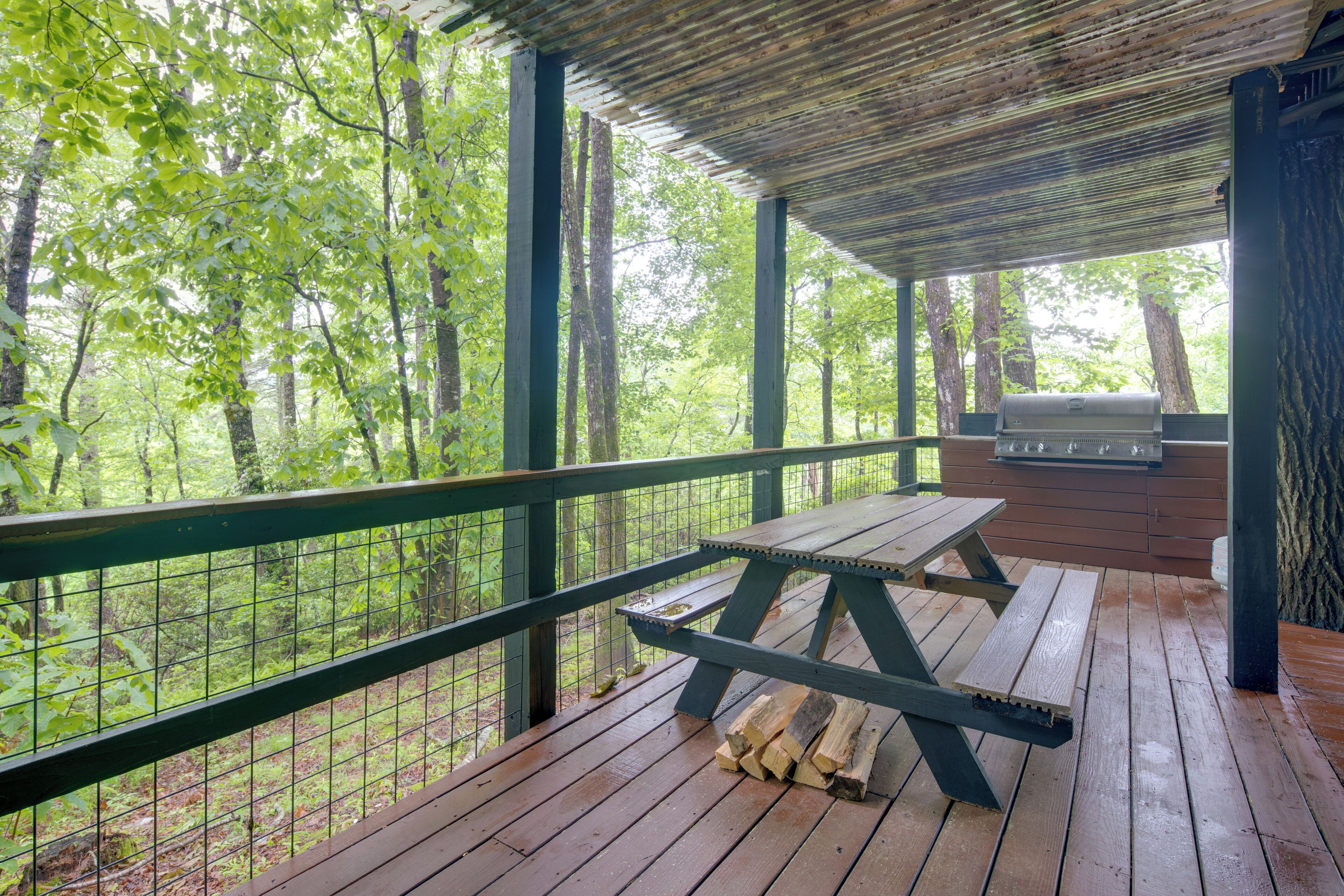 Eine überdachte Holzterrasse mit Picknicktisch und Grill, umgeben von Bäumen.