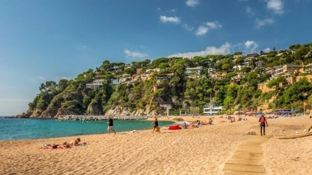 Une plage de sable avec une promenade en bois, des gens qui prennent des bains de soleil et, au loin, des maisons sur une colline.