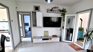 Un salon moderne avec un téléviseur à écran plat, un pouf gris et un coin repas visible à travers une porte ouverte.