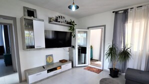 Un salon moderne avec un téléviseur à écran plat, un canapé gris, un pouf rond et une plante en pot.