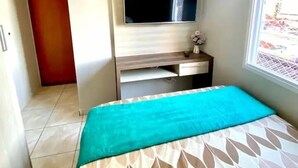 A hotel room with a bed, a TV mounted on the wall, a desk with a chair, and a window providing a view of the outside. - Apto Boqueirão próximo ao Mar (Praia Grande)